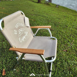 Beige camping chair with wooden armrests on grass near a body of water