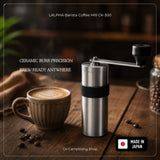 LALPHA Barista Coffee Mill CK-300 manual coffee grinder with a stainless steel body, black ceramic burr, and black handle, displayed next to a cup of latte on a wooden table with coffee beans.