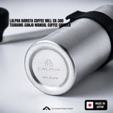 Close-up of a brushed silver cylindrical coffee grinder body with a black strap, shown on a light gray background with text reading 'LALPHA BARISTA COFFEE MILL CK-300 TSUBAME-SANJO MANUAL COFFEE GRINDER' and a 'MADE IN JAPAN' badge.