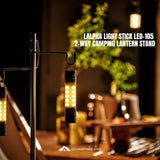 A LALPHA LIGHT STICK LED-105 2-WAY CAMPING LANTERN STAND shown lit up in a warm, cozy camping setting with a blurred background of a campfire and green foliage. The product features a black metal stand with two hanging cylindrical LED lanterns emitting a warm yellow light.