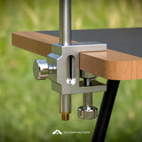 A close-up of a silver metal clamp mount attached to a wooden table leg, with a vertical metal post extending upward. The background is a blurred green outdoor setting, and a 'GO CAMPTHING SHOP' logo watermark is visible at the bottom.