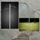 A metal hanging medical IV pole shown in two views: a studio-style shot on a dark background and a smaller outdoor photo mounted on a textured gray wall. The outdoor inset shows the pole inserted into grass.