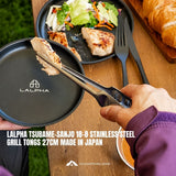 A hand uses stainless steel tongs to lift a grilled piece of chicken from a black LALPHA plate. The scene is set on a wooden table with a salad, a fork, and a croissant in the background, with text overlays describing the LALPHA Tsubame Sanjo 18-B stainless steel grill tongs.