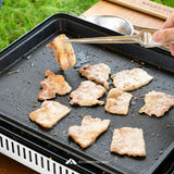 Grilled pork slices cook on a black outdoor camping griddle, with a hand using metal tongs to flip a piece. A wooden cutting board, metal bowl, and grass are visible in the background.