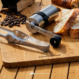 A stainless steel manual coffee grinder and matching metal tongs with LALPHA branding, styled on a wooden cutting board with coffee beans, sliced bread, and a bag of coffee in a warm outdoor setting.