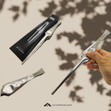 A hand holds a silver aluminum camping spoon against a beige background with leaf shadows, alongside its black packaging and a close-up of the spoon handle. The brand 'Lalique' is visible on the packaging and handle.