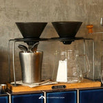 Coffee-making setup with coffee filters, a metal cup, and a glass pitcher on a wooden surface.