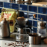 Coffee-making setup with Hills Bros coffee bag, grinder, and beans on a blue tiled wall background.