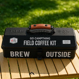 A black metal field coffee kit box with a brown leather handle sits on a wooden slatted table outdoors, with blurred green grass in the background. The box is labeled 'TOOL BOX', 'GO CAMPTHING FIELD COFFEE KIT', and 'BREW OUTSIDE'.